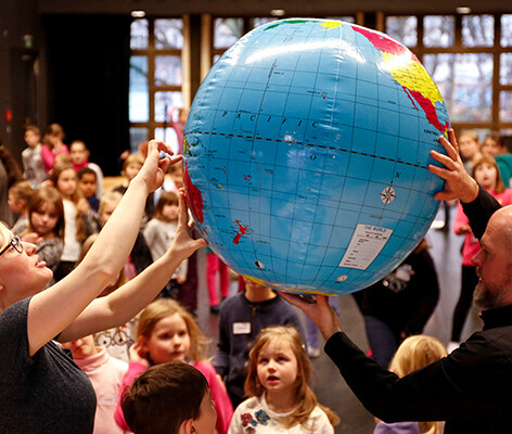 Zwei Musiktheater-Pädagog*innen zeigen einer staunenden Gruppe Kinder Orte auf einer großen aufblasbaren Weltkugel