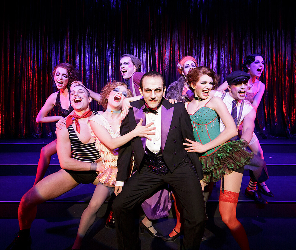 Szene aus "Cabaret": Ein Sänger im Anzug wird im Halbkreis von 8 weiteren Sänger*innen umringt, alle schauen in den Zuschauerraum