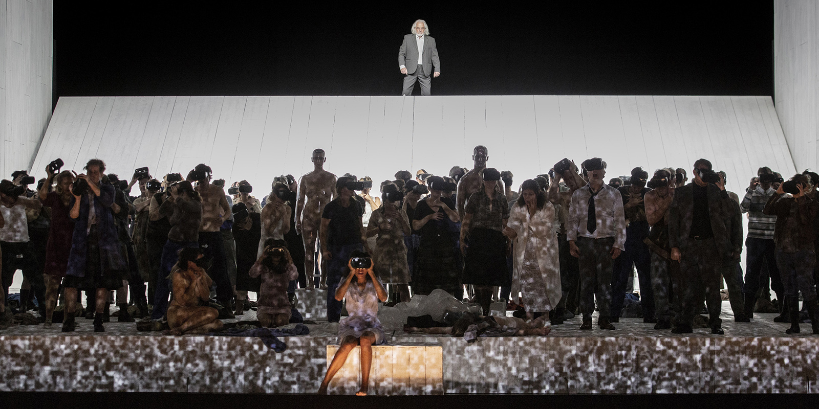 Szene aus "Moses und Aron": Viele Sänger*innen stehen verteilt vor einem einzelnen Sänger, der erhöht im hinteren Teil der Bühne stehtsteht